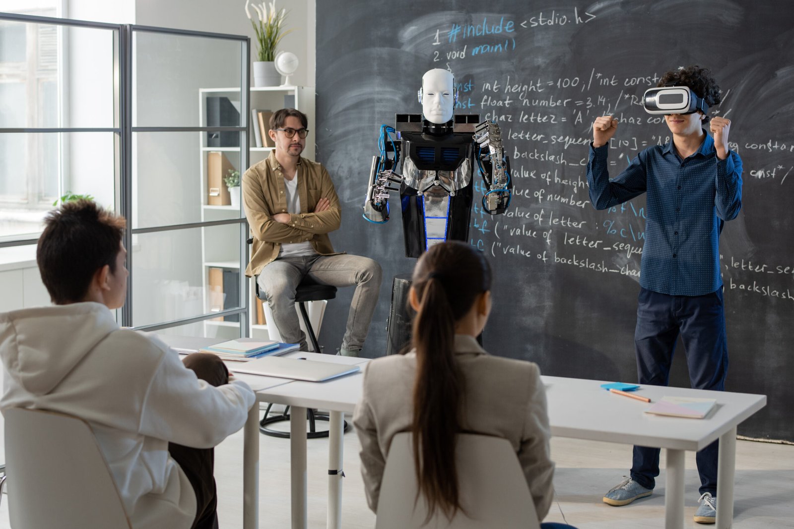 Teenage student with vr headset standing by blackboard in front of his classmates and demonstrating abilities of robot near by